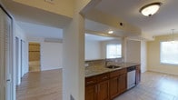 This single-story, one-bedroom, one-bathroom residence offers a thoughtfully designed layout that balances functionality and comfort. The kitchen serves as a central feature of the home, showcasing warm medium-toned wood cabinetry paired with granite countertops that provide generous prep space. A stainless steel sink and dishwasher add a modern, practical touch, while a classic tile backsplash introduces subtle texture to the space. The kitchen opens toward the adjacent living area through a structural column framing, creating a sense of visual connectivity between the two spaces. Light tile flooring runs through the kitchen, transitioning to warm hardwood-style flooring in the living areas, adding a cohesive flow throughout the home. Recessed lighting provides even illumination in the kitchen, while large windows dressed with vertical blinds allow natural light to filter into the living space. The neutral, light-toned walls throughout contribute to a bright and airy ambiance. Overall, the residence presents a clean, move-in-ready aesthetic with durable finishes well-suited for everyday living.