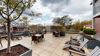 Located at 927 Drake St in Madison, Wisconsin, this property showcases thoughtfully designed outdoor common spaces that blend urban architecture with comfortable open-air living. The building's exterior combines classic red brick with modern panel cladding in warm ochre and neutral tones, creating a contemporary yet inviting aesthetic.

The ground-level courtyard serves as a welcoming transitional space, featuring large raised planters filled with ornamental shrubs and small trees set against a backdrop of gravel landscaping. Wide-format tile pavers form a clean walkway leading to a glass-paneled entrance door, while metal railings and wood-beamed overhangs add architectural warmth. Balconies overlooking the courtyard reinforce the building's multi-level design.

The rooftop terrace is a standout feature, offering an expansive open-air gathering space finished with large neutral tiles. Multiple seating arrangements — including dining table sets with chairs and umbrella shade, lounge chairs, and Adirondack-style seating — provide versatile options for relaxation and socializing. A gas grill station enhances the space's outdoor entertaining capabilities. Surrounded by metal railings, the terrace offers open views of Madison's surrounding neighborhood rooftops and tree canopy beneath an open sky.