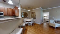 This single-floor residence offers a thoughtfully designed layout with two bedrooms and two bathrooms across a comfortable footprint. The open-concept living, dining, and kitchen area forms the heart of the home, connected by warm hardwood-style flooring and accented with crown molding throughout. The kitchen features rich medium-toned wood cabinetry, stainless steel appliances, granite-style countertops, and a raised breakfast bar with pendant lighting, opening directly to the dining area. The dining space accommodates a full table setting and is enhanced by decorative wall accents and a ceiling-mounted light fixture.

The living room benefits from natural light via large windows and a glass door leading to an outdoor area, complemented by a ceiling fan. A hallway connects the main living area to the private quarters. The primary bedroom includes carpeted flooring, a window with outdoor views, and a walk-in closet with wire shelving. The second bedroom similarly features carpeted floors and closet access. Both bathrooms are finished with tile flooring, warm wood vanities, and large mirrors. A dedicated walk-in closet with carpet flooring and wire shelving provides additional storage.