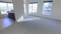 This single-floor space presents a clean, modern interior characterized by an open layout and an abundance of natural light. Large windows along the exterior walls frame views of the surrounding urban landscape, including nearby buildings and mature trees, creating a strong visual connection to the outdoors. The windows allow sunlight to pour in throughout the day, casting warm patches of light across the neutral-toned carpet that covers the main living area.

The walls are finished in crisp white, contributing to a bright and airy atmosphere. A rounded structural column serves as a subtle architectural detail, adding a sense of design intention to the otherwise minimalist space. The open-plan configuration allows for flexible furniture arrangement and a free-flowing sense of space. Toward one side, dark cabinetry is partially visible, suggesting the presence of a kitchen or storage area that anchors the layout.

Overall, the space conveys a contemporary, uncluttered aesthetic with practical finishes. The combination of ample glazing, neutral flooring, and clean architectural lines makes it a versatile environment well-suited to a variety of uses.
