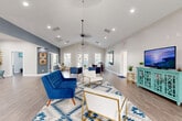Located at 1069 Chateau Crossing Dr in Fort Mill, SC, this single-floor community space offers a well-designed layout that seamlessly blends comfort and functionality. The main living area is defined by soaring vaulted ceilings with recessed lighting and a ceiling fan, creating an airy and open atmosphere. Large French doors and multiple windows flood the space with natural light while providing views of the surrounding community. Light hardwood-style flooring runs throughout, complementing the neutral gray and white color palette accented by bold slate-blue feature walls.

The living area is thoughtfully arranged with distinct seating zones, including plush royal blue velvet sofas and gold-framed accent chairs arranged over a geometric area rug, alongside a dedicated workspace with a desk and office area. A teal media console with a flat-screen TV anchors one end of the room, while a reception-style pass-through window and private office nook add a professional touch.

The kitchen is equipped with rich dark espresso cabinetry, granite countertops, stainless steel appliances including a dishwasher, range, and microwave, and durable wood-look flooring. A café-style corner with decorative signage adds a welcoming finishing detail to this well-appointed space.