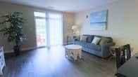 This single-floor space offers a welcoming and comfortable layout designed with a relaxed, coastal aesthetic. The living area serves as the heart of the space, showcasing smooth hardwood-style flooring that adds warmth and a clean, modern feel underfoot. A large sliding glass door floods the room with natural light, framing a view of the lush greenery outside while providing easy access to a private patio area, where outdoor seating invites relaxation. Sheer vertical blinds softly diffuse the incoming light, creating a bright yet comfortable atmosphere throughout the day. The living space is thoughtfully appointed with a plush gray sofa accented by patterned and solid throw pillows, complemented by an ornately carved octagonal coffee table that adds an artistic focal point. A sleek console table with a classic table lamp provides additional functionality and a touch of elegance. Completing the coastal ambiance, a large canvas painting of a serene beach scene adorns the wall, tying the interior design together with a tranquil, seaside-inspired theme. The overall atmosphere is airy, inviting, and well-suited for comfortable everyday living.
