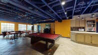 This single-floor recreational and social space makes an immediate visual impact through its bold design choices. The most distinctive architectural feature is the fully exposed structural ceiling, painted a deep cobalt blue, which reveals beams, joists, and mechanical systems overhead — lending the space an industrial yet energetic character. Recessed and track lighting is integrated throughout, ensuring the interior is well-lit despite the dark overhead palette.

The walls are finished in a warm, saturated amber-gold tone, creating a lively contrast against the blue ceiling and accents. Large multi-pane windows along one wall flood the space with natural light and provide views of the surrounding greenery outdoors.

The main open area is anchored by a full-size billiard table with a deep red felt surface, positioned centrally on tile flooring. Adjacent to this is a dining and lounge area furnished with round and rectangular tables and wooden chairs, suitable for group gatherings. An upright piano rests against the amber wall alongside framed artwork, adding a cultural and social dimension to the space.

A dedicated kitchenette offers light oak cabinetry, a sink, tile countertops, and a stack of appliances including microwaves and a warming oven — well-suited for event catering or everyday use. Blue-painted doors and trim tie the color scheme together throughout, reinforcing the cohesive and intentional design of this engaging communal space.