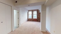 This single-floor unit showcases a compelling mix of historic character and contemporary convenience. The standout feature of the main living area is a full exposed brick accent wall, framed by two large windows that draw in generous natural light and lend the space an unmistakable urban loft aesthetic. Soft, neutral carpeting covers the living area floor, contributing to a warm and comfortable ambiance. Structural columns and defined ceiling soffits add architectural interest, while clean white walls provide a crisp, versatile backdrop throughout.

The open layout allows for a seamless visual connection between the living area and the kitchen, which is accessible via a pass-through counter. The galley-style kitchen is efficiently designed, featuring ample cabinetry with wood-trimmed accents, light-toned countertops, and black appliances including a coil-top electric range, a built-in microwave, and a dishwasher. Recessed track lighting illuminates the workspace, and warm hardwood flooring grounds the kitchen area with a rich contrast to the rest of the unit.

The bathroom is clean and functional, offering a full tub-shower combination with white tile surround, a toilet, and a vanity with a countertop sink and mirror. Overall, the space presents a cohesive blend of industrial heritage and practical modern living.
