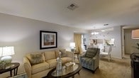 This single-floor residence offers a thoughtfully designed open-concept layout that seamlessly connects the living and dining areas, creating a sense of spaciousness and flow. The living area is appointed with a plush cream sofa, a patterned accent chair, and a glass-topped coffee table, all set atop neutral carpeting that runs throughout the space. Warm, soft lighting from a classic table lamp complements the natural light filtering through sheer gold drapery, lending the room a cozy yet refined ambiance. A framed piece of abstract artwork adorns the wall, adding a tasteful decorative accent. Transitioning into the dining area, a chandelier with elegant globe lights anchors the space above a formal dining table surrounded by white upholstered chairs, creating an inviting setting for entertaining. A glimpse into what appears to be a bright kitchen is visible beyond, suggesting a well-connected and functional floor plan. The overall palette of soft creams, taupes, and warm golds gives the entire residence a cohesive, welcoming aesthetic that balances comfort with classic style.