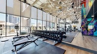 Located in the heart of downtown Phoenix at 802 N 1st Ave, this two-story fitness facility offers a comprehensive and visually dynamic workout environment. The larger main floor is anchored by soaring ceilings and expansive floor-to-ceiling windows that frame sweeping views of the Phoenix skyline, flooding the space with abundant natural light. This level is outfitted with a wide array of strength and conditioning equipment, including free weights, cable machines, and plate-loaded stations, all set against bold, vibrant murals that energize the atmosphere. A sculptural, geometric ceiling installation adds a striking artistic dimension to the workout floor. The upper floor continues the fitness experience with cardio equipment — including treadmills, cycling bikes, and ellipticals — positioned directly in front of glass walls overlooking a pool terrace and the surrounding cityscape. A TRX and functional training zone, complete with a heavy bag, rounds out the upper level. Connecting the floors are two staircases, while a clean, neutral-toned hallway leads to the wellness amenities: two beautifully appointed cedar saunas with herringbone wood flooring and tiered benching, as well as two bathrooms finished with light tile and modern fixtures. The facility balances high-energy design with refined recovery spaces throughout.