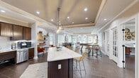 Located at 7100 Claxton Cir in Raleigh, NC, this thoughtfully designed single-floor community space offers a generous and versatile layout ideal for gathering and recreation. The interior is unified by rich wood-look tile flooring, tray ceilings with recessed lighting, and neutral warm-toned walls that lend a polished, welcoming ambiance throughout.

At the heart of the space is an open-concept kitchen and social area featuring dark espresso cabinetry, white quartz countertops, stainless steel appliances, and a large central island with seating for multiple guests. Floating wood shelves add a decorative yet functional touch nearby.

The lounge area is anchored by a classic stone fireplace with a substantial mantle, a wall-mounted television, and an abundance of natural light streaming through multiple white-framed windows that frame lush outdoor views. Elegant tray ceilings add architectural depth to this relaxing retreat.

A dedicated game room provides space for recreation, complete with a foosball table, bistro-style seating, and direct access via a set of glass-paned French doors to a private sitting room furnished with soft lounge chairs — perfect for quieter gatherings or meetings. Additional seating nooks throughout the space offer intimate areas for conversation, all bathed in natural light from generously sized windows.