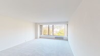 This single-floor unit offers a clean, neutral aesthetic throughout, with white walls and smooth ceilings that create a bright and airy atmosphere. The spacious living area is a standout feature, anchored by a wide, multi-panel window that spans a large portion of the exterior wall, inviting abundant natural light and framing scenic views of surrounding treetops and the urban skyline beyond. Soft, plush wall-to-wall carpeting runs throughout the living and sleeping areas, contributing to a comfortable and cohesive feel. A second carpeted room, likely a bedroom, also benefits from natural light through its own window, with views of neighboring buildings providing a sense of urban connectivity. The galley-style kitchen is efficiently laid out with white cabinetry lining both sides, dark granite-style countertops, a stainless steel sink, a dishwasher, and a full-size refrigerator, all set on light vinyl flooring. The bathroom features a vanity with warm wood cabinetry, a light countertop, a large wall mirror with overhead lighting, and classic tile flooring with a subtle diamond pattern. The overall layout, as seen in the overhead dollhouse view, is compact yet well-organized, with a clear flow between the living, sleeping, kitchen, and bathroom areas.