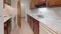 This single-floor unit offers a straightforward and functional layout suited for everyday living. The kitchen is equipped with dark wood cabinetry featuring brushed nickel hardware, light-toned laminate countertops, a stainless steel double-basin sink, and white appliances including a range and dishwasher. Light wood-look vinyl flooring runs throughout the kitchen, providing a clean and durable surface.

The living area is generously sized, finished with neutral carpet and neutral-toned walls. A large sliding glass door with vertical blinds allows natural light to fill the space. An adjacent dining area is highlighted by a simple chandelier and connects visibly to the kitchen, where a refrigerator is accessible nearby. A wood-paneled closet is positioned conveniently off this shared living-dining space.

The bedroom features plush carpet, a window with horizontal blinds admitting natural light, and a spacious reach-in closet with bi-fold wood doors and an upper storage shelf. The bathroom is finished with light wood-look vinyl flooring, a grey vanity with a white countertop, a full-size tub and shower combination with a white surround, and a curved shower rod. Neutral wall colors are consistent throughout the unit, contributing to a cohesive and understated overall ambiance.