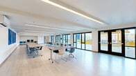 Located at 916 N 143rd St in Seattle, WA, this single-floor multipurpose room offers a generous, open layout well-suited for a variety of uses including community gatherings, instructional sessions, and collaborative events. The space features light hardwood-style flooring throughout, creating a clean and welcoming atmosphere that is further enhanced by crisp white walls and a modern, minimalist aesthetic.

Overhead, linear LED lighting runs along the ceiling in parallel rows, providing bright, even illumination across the entire room. The ceiling itself features subtle architectural detailing with recessed tray elements that add visual depth. A ceiling-mounted projector and a wall-mounted split HVAC unit ensure the room is equipped for both presentations and year-round comfort.

One of the room's most notable features is the extensive glazing along one wall, comprising large sliding glass panels and a set of double doors that open directly to an outdoor patio area, flooding the interior with natural light and offering seamless indoor-outdoor connectivity. Views of the surrounding exterior space are visible through the full-height glass.

The room is furnished with mobile tables and multi-colored chairs arranged in flexible configurations, making it easy to adapt the layout for different group sizes or activities. Decorative blue artwork panels on one wall add a distinctive visual accent. A computer station and additional storage are visible along one end of the room.