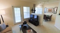 This single-story space showcases a thoughtfully designed open-concept layout that seamlessly connects the living and dining areas. The living room is anchored by plush wall-to-wall carpeting and features a sleek media console with an integrated X-pattern storage unit, providing both functional storage and a stylish focal point for the flat-screen television displayed above. Track lighting runs along the ceiling, supplementing the generous natural light that streams in through multiple large windows fitted with horizontal blinds, including a full-length glass door that opens to an outdoor area. Warm neutral tones on the walls and crown molding details lend a clean, polished aesthetic throughout. A half-wall partition with a flat ledge serves as a natural divider between the living space and the adjacent dining area, where dark wood dining chairs are visible. Decorative accents, including tall black vase-style ornaments atop the partition and a globe centerpiece on the dark wood coffee table, add character and sophistication. The overall ambiance is bright, open, and welcoming, with a cohesive design palette that balances comfort and style.