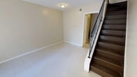 This two-story space offers a straightforward and functional layout designed for practical everyday living. The main level features an open living area finished with light-toned tile flooring that reflects ambient light, creating a bright and airy atmosphere. The walls are painted in a neutral cream hue throughout, lending the space a clean and cohesive aesthetic that serves as a versatile backdrop for a variety of furnishing styles. A flush-mount ceiling light provides overhead illumination, while a visible air vent indicates a central HVAC system for year-round climate control. A doorway from the main living area leads to an adjoining room, hinting at additional functional spaces on the ground floor. The staircase is a notable architectural feature, constructed with dark wood treads that provide a warm contrast against the light surroundings, and fitted with sleek metal balusters for a contemporary touch. White-painted baseboards frame the rooms consistently, adding a polished finishing detail throughout. The second floor expands the usable area of the home, accessible via the centrally positioned staircase. Overall, this space presents a clean, move-in ready environment with a neutral palette and classic design elements suited to a wide range of uses.