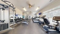 Located at 130 Canary Pl in Burlington, NC, this single-floor fitness center offers a comprehensive workout facility thoughtfully divided into two distinct zones. The main exercise area features durable rubber flooring and is outfitted with an extensive array of cardio equipment, including treadmills, ellipticals, stationary bikes, and stair climbers. A dedicated free weights section provides a full rack of dumbbells alongside adjustable benches, while a multi-station cable machine supports a wide range of strength training exercises. Multiple wall-mounted flat-screen televisions are positioned throughout the space for entertainment during workouts. Ceiling fans and recessed lighting keep the environment comfortable and well-lit, while crown molding lends a refined architectural touch to the interior. Large windows with blinds allow natural light to fill the room while offering views of the surrounding greenery. A separate studio room, accessible through a doorway, features light hardwood-style flooring, floor-to-ceiling mirrors, a wall-mounted television, and a storage unit for accessories such as medicine balls. This studio is ideal for yoga, stretching, or group fitness activities. French doors in the main area provide additional access and contribute to the bright, open ambiance of this well-appointed facility.
