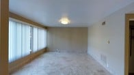 This single-floor space presents a clean, functional layout finished with consistent design elements throughout. The main living area is characterized by light-toned marble-style tile flooring that extends seamlessly across the space, lending a polished and easy-to-maintain surface underfoot. Walls are finished in a warm neutral beige tone, creating a versatile backdrop suitable for a variety of interior styles.

Natural light is a notable feature, with large windows dressed in vertical blinds allowing residents to control light levels while maintaining privacy. Flush-mount ceiling light fixtures provide additional ambient illumination. The smooth white ceilings contribute to a clean, open feel throughout.

Visible from one vantage point, cabinetry with warm wood tones and what appears to be stone countertops suggest a kitchen area with practical storage and workspace. A white interior door is also visible, indicating clear separation between living zones. HVAC vents are present along the walls and floors, ensuring climate control throughout the space. Overall, the interior offers a straightforward, move-in-ready environment with neutral finishes and ample natural light.