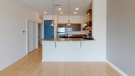 This well-designed single-floor residence offers a clean, modern aesthetic throughout. The open-concept kitchen serves as a central highlight, outfitted with stainless steel appliances including a refrigerator, range with a stainless steel hood vent, and dishwasher. Rich dark wood cabinetry pairs elegantly with granite countertops, while a breakfast bar with pendant lighting creates a natural gathering point. Light hardwood flooring extends seamlessly through the kitchen and into the living area, which is bright and open with large sliding glass doors that invite abundant natural light and provide views of the surrounding neighborhood. The bedroom is finished with soft carpeting, a ceiling fan, and a sliding window that frames outdoor scenery. The bathroom impresses with a long granite-topped vanity featuring vessel sinks, dark wood cabinetry, a large illuminated mirror, and floor-to-ceiling dark tile surrounding the soaking tub and shower area. Tile flooring continues into a dedicated walk-in closet, which offers dual clothing rods and generous shelving on both sides, providing exceptional storage capacity. The overall ambiance of the space is modern, clean, and functional, with thoughtful finishes and a neutral color palette throughout.