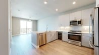 This thoughtfully designed one-bedroom, two-bathroom residence offers a smart, efficient layout across a single floor. The open-concept kitchen and living area serves as the heart of the home, featuring light hardwood-style flooring, recessed lighting, and a ceiling fan. The kitchen is outfitted with a full suite of stainless steel appliances — including a gas range, built-in microwave, dishwasher, refrigerator, and a dedicated wine cooler — complemented by white cabinetry, a classic subway tile backsplash, and light countertops with a functional peninsula. Large sliding glass doors open to a private balcony, flooding the living area with natural light and offering open outdoor views. The bedroom is carpeted for comfort, featuring large windows that frame expansive views, a ceiling fan, and direct access to a walk-in closet with built-in shelving and hanging rods on multiple sides. A conveniently located hallway houses a stacked in-unit washer and dryer. Two bathrooms provide added convenience for residents and guests alike. The overall ambiance is clean, modern, and bright, with a neutral palette throughout that creates a calm and cohesive living environment.