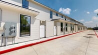This exterior presents a contemporary multi-unit building with a clean, modern farmhouse aesthetic. The facade is finished in crisp white board-and-batten siding, accented by bold black-framed windows that provide strong visual contrast and ample natural light to the units within. Each unit features its own private gated entry with white vertical-panel gates, offering individual access and a degree of privacy for each occupant.

The building stretches along a well-maintained concrete drive that serves both as a parking area and a fire lane, clearly delineated by a bold red curb stripe and painted white stall markings. The lot provides generous parking capacity directly adjacent to the unit entries, ensuring convenient access for residents and visitors alike.

The surrounding landscape offers a natural counterpoint to the building's modern design. Established mature trees, including large oaks, frame the property and provide shade, while native shrubs and greenery line the perimeter fence. A wood privacy fence along one boundary adds additional separation from neighboring properties.

The property benefits from an open, sunny exposure beneath a broad Texas sky, giving the exterior a bright and welcoming character. The combination of new construction detailing, practical site planning, and lush mature landscaping creates a functional yet visually appealing environment that balances contemporary design with its natural surroundings.