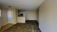 This single-floor space features an open-concept layout that connects the living area to the kitchen, creating a practical and functional flow. The living area is finished with dark wall-to-wall carpeting and neutral-toned walls, offering a understated backdrop adaptable to various furnishing styles. A ceiling fan is centrally positioned, providing both air circulation and modest overhead lighting to the space.

The kitchen is defined by a white peninsula that serves as a natural divider between the cooking and living areas. White cabinetry lines the kitchen walls, complemented by a built-in microwave and a full-size refrigerator. Fluorescent overhead lighting is installed beneath the upper cabinetry, illuminating the kitchen workspace effectively.

The textured popcorn ceiling is consistent throughout the space, adding a classic residential character. Neutral baseboards frame the carpeted floors cleanly. An entryway is visible to one side, hinting at additional rooms beyond the main living area. Overall, the space presents a straightforward, functional layout suited for everyday living.