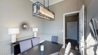 This single-floor space showcases a thoughtfully designed interior that blends classic architectural charm with contemporary style. The dining area is a standout feature, highlighted by a striking rectangular pendant light fixture with a dark metal geometric frame and frosted panel shades, which serves as a dramatic focal point above the dark dining table. Upholstered dining chairs in a soft blue-grey tone add a modern, sophisticated touch to the setting. The walls are finished in a neutral warm grey palette and accented with classic wainscoting — crisp white paneling that runs along the lower portion of the walls — lending the space a timeless, polished character. Crown molding at the ceiling line further reinforces the refined architectural detailing throughout. A decorative starburst wall mirror adds an artistic element to the dining space, complementing the contemporary aesthetic. Dark hardwood flooring grounds the room with warmth and depth, visible through the doorway leading to an adjacent area. The overall ambiance is welcoming and elegant, balancing traditional architectural elements with modern furnishings and fixtures to create a cohesive and well-appointed living environment.