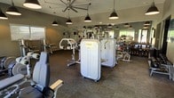 This fitness center offers a thoughtfully designed workout environment spread across a single floor. The space is outfitted with a robust collection of professional-grade strength training machines, including cable systems, weight stack machines, and a dedicated free weights area with a full dumbbell rack. Large mirrors line the walls, amplifying the sense of space and allowing users to monitor their form throughout their workouts. The flooring features durable, dark carpet tiles suited for high-traffic athletic use. Overhead, a combination of industrial-style black pendant lights and a large ceiling fan ensures the space remains well-lit and comfortably ventilated. Natural light filters in through windows along the perimeter, adding brightness to the interior. The muted green-gray wall tones contribute to a focused and calm workout atmosphere. The layout is open and organized, providing ample room to move between equipment. Overall, the fitness center balances functionality with a clean, modern aesthetic, making it a practical and inviting space for a full range of strength and conditioning workouts.