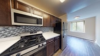 This single-floor residence offers a well-designed layout that balances functional living spaces with natural light. The kitchen is a standout feature, showcasing rich dark-brown cabinetry with brushed nickel hardware, white countertops, and a decorative mosaic tile backsplash in shades of gray and blue. Stainless steel appliances include a gas range, over-the-range microwave, and refrigerator, providing a cohesive, modern aesthetic.

The living areas are finished with dark hardwood-style flooring that adds warmth and contrast to the neutral walls. Generously sized windows frame views of surrounding trees, allowing abundant natural light to fill the space. A baseboard heating unit runs beneath the windows in both the living area and bedrooms, ensuring year-round comfort.

The bedrooms are carpeted in a neutral tone and feature multiple windows with wooded views, built-in closets with bifold doors, and ample room for furnishings. The bathroom is finished with white tile wainscoting, a tub-shower combination, a white vanity with storage, a wall-mounted mirror, and tile flooring, creating a clean, classic appearance throughout.