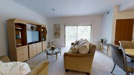 This single-floor space showcases a welcoming living room designed for comfort and functionality. The room is finished with plush wall-to-wall carpeting in a neutral tone, complementing the light-colored walls that enhance the sense of openness. A large sliding glass door fitted with vertical blinds serves as a prominent architectural feature, filling the space with abundant natural light while offering direct access to an outdoor area with greenery visible beyond. The living area is centered around a comfortable upholstered sofa and matching armchair in warm golden tones, paired with a glass-top coffee table that adds a touch of elegance. A built-in or freestanding wooden entertainment unit with a flat-screen TV, shelving, and drawer storage occupies one wall, providing both practical storage and a focal point for the room. Decorative accents, including wall art and small tabletop accessories, contribute to the inviting atmosphere. The overall ambiance is bright, clean, and livable, making effective use of the available space while maintaining a relaxed and comfortable aesthetic.