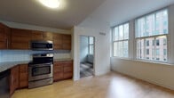 This single-floor residence is organized into a well-defined layout comprising a kitchen, living area, bedroom, and bathroom. The kitchen is a standout feature, outfitted with warm wood cabinetry, granite countertops, a white tile backsplash, and a full suite of stainless steel appliances including a double oven range, microwave, dishwasher, and refrigerator. Light hardwood flooring runs through the kitchen and living area, creating a cohesive and bright aesthetic. Large windows in the living area and bedroom allow natural light to fill the space and offer views of the surrounding urban environment. The bedroom is carpeted and features access to the rest of the unit through a standard interior door, with ceiling-height walls that contribute to an airy feel. The bathroom is finished with large-format neutral tile flooring, a bathtub and shower combination, a wall-mounted towel bar, and a wood-accented vanity with a white countertop and mirror cabinet. Clean white walls and simple, modern fixtures throughout the unit contribute to a consistent, understated aesthetic.