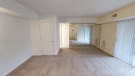 This single-floor residential space presents a clean and functional layout designed with comfort and practicality in mind. The interior is finished with wall-to-wall plush carpeting in a warm neutral tone, providing a soft and cohesive look across the living areas. The walls are painted in a soft off-white, contributing to an overall light and airy ambiance that makes the space feel open and welcoming.

A standout feature of the main room is a large floor-to-ceiling mirrored wall, which spans a significant portion of one wall and serves a dual purpose — functioning as closet doors while visually expanding the room and amplifying natural light. The mirrored panels are framed in gold-toned trim, adding a subtle classic touch to the interior design.

The space includes multiple paneled doors in crisp white, adding a traditional architectural detail that complements the neutral palette. A hallway visible through the main room provides a sense of depth and connectivity between spaces, hinting at additional rooms and functional areas beyond. A wall-mounted thermostat or intercom panel is also visible, pointing to the unit's practical amenities. Overall, the space offers a bright, well-maintained environment with timeless design elements suited for comfortable everyday living.
