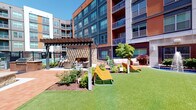 This contemporary residential community is designed to offer an inviting blend of indoor comfort and outdoor living. The building's exterior showcases a striking mix of dark blue siding, warm brick red paneling, and large windows, with private balconies that overlook the meticulously maintained grounds below.

At the heart of the community lies a vibrant courtyard, thoughtfully landscaped with lush green lawn, decorative red Japanese maples, flowering shrubs, and mulched garden beds. A central fountain serves as a focal point, surrounded by multiple seating arrangements with plush outdoor sofas and modern lounge chairs, creating an ideal setting for relaxation and socializing. A handsome pergola structure with a stone accent wall anchors one corner of the courtyard, offering a shaded gathering space adjacent to a pool area enclosed by black iron fencing.

The building entrance is marked by floor-to-ceiling glass walls and double glass doors, flanked by large potted plants and warm brick pavers that set a welcoming tone. Interior finishes are clean and neutral, featuring crisp white walls, panel-style doors with brushed nickel hardware, and tile flooring. The walk-in closet offers generous storage capacity with wire shelving systems that include multiple tiers for folded items, overhead hanging rods, and upper basket shelving, maximizing organizational potential throughout.