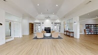 Located at 1925 Ashland City Rd in Clarksville, TN, this single-floor community space offers a refined and welcoming atmosphere across a generously sized layout. Light hardwood flooring runs throughout, creating a cohesive and warm foundation for each distinct area.

The main living room is an elegant, open space anchored by a classic fireplace and a soaring vaulted ceiling that enhances the sense of grandeur. A statement crystal chandelier hangs overhead, illuminating a seating arrangement of plush cream armchairs around a marble coffee table, all set atop a bold black-and-white patterned rug. Large glass-paneled doors line the perimeter, flooding the room with natural light and providing views of the outdoors.

A second living room offers a more relaxed, casual atmosphere with deep blue-gray accent walls, comfortable upholstered sofas, a dramatic wrought-iron candle chandelier, and direct access to an outdoor patio area. A built-in chalkboard wall adds a unique, interactive element to the space.

The dining room is a sophisticated setting featuring a large marble-topped table surrounded by clear acrylic chairs, a dalmatian-print area rug, and a cascading sculptural chandelier. Bold abstract artwork and tall windows contribute to its contemporary character.

The hallway serves as a bright, inviting entry point with bay-style windows, elegant seating, and a full-length mirror, seamlessly connecting all areas of this well-appointed space.