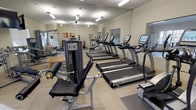 This fitness center is thoughtfully designed to support a full range of workout routines within a single-level layout. The space is organized into distinct functional zones, each catering to different aspects of physical training. Large windows along one wall allow natural light to fill the room, offering views of the surrounding exterior while keeping the environment bright and welcoming.

The cardio zone features a row of TRUE-brand treadmills alongside elliptical machines, all equipped with individual digital displays for tracking workout metrics. A ceiling fan and overhead fluorescent lighting keep the space comfortable during use. Full-length mirrors line the walls, visually expanding the room and allowing users to monitor their form.

The strength training area includes a multi-functional cable machine and a leg extension/curl unit, providing options for both upper and lower body resistance work. Wooden benches are positioned nearby for stretching or rest. A mounted flat-screen television adds an entertainment option for those using cardio equipment.

A dedicated free weights and functional training corner houses a full rack of dumbbells in varying weights, a medicine ball tower, stability balls, and a spin bike. Colorful artwork adorns the walls, and rubber matting underfoot provides safety and cushioning. The overall ambiance is clean, practical, and well-maintained.
