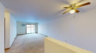 This single-floor residential unit offers a practical and functional layout across several well-defined areas. The spacious living room is a standout feature, boasting wall-to-wall carpeting, a ceiling fan with a built-in light fixture, baseboard heating, and a wall-mounted air conditioning unit. A sliding glass door floods the space with natural light and provides direct access to an outdoor patio area. A low half-wall partition subtly defines the transition between spaces.

The kitchen is equipped with ample white cabinetry framed in warm wood trim, generous countertop space, a double stainless steel sink, and a dishwasher, providing a clean and organized cooking environment.

The bedrooms are carpeted and well-lit by large windows that offer outdoor views. Each room features baseboard heating and wall-mounted AC units for year-round climate control. One bedroom includes a sliding wood-panel closet, adding practical storage. The neutral white walls throughout lend a clean, bright feel to every room.

The bathroom is efficiently designed with a vanity sink, toilet, a large mirror with overhead lighting, and warm wood-trimmed door accents that tie into the overall aesthetic of the unit.