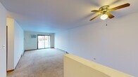 This single-floor residential unit offers a practical and functional layout across several well-defined areas. The spacious living room is a standout feature, boasting wall-to-wall carpeting, a ceiling fan with a built-in light fixture, baseboard heating, and a wall-mounted air conditioning unit. A sliding glass door floods the space with natural light and provides direct access to an outdoor patio area. A low half-wall partition subtly defines the transition between spaces.

The kitchen is equipped with ample white cabinetry framed in warm wood trim, generous countertop space, a double stainless steel sink, and a dishwasher, providing a clean and organized cooking environment.

The bedrooms are carpeted and well-lit by large windows that offer outdoor views. Each room features baseboard heating and wall-mounted AC units for year-round climate control. One bedroom includes a sliding wood-panel closet, adding practical storage. The neutral white walls throughout lend a clean, bright feel to every room.

The bathroom is efficiently designed with a vanity sink, toilet, a large mirror with overhead lighting, and warm wood-trimmed door accents that tie into the overall aesthetic of the unit.