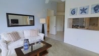This single-floor space showcases an airy, coastal-inspired design highlighted by notably high vaulted ceilings that lend a sense of openness and grandeur to the living area. The main living space is finished with neutral-toned carpet and painted in soft, calming blue-grey tones that reinforce a relaxed, beach-inspired ambiance. A ceiling fan is mounted overhead, serving both a functional and aesthetic purpose within the tall ceiling space.

The living area centers around a glass-topped coffee table and a white sofa accented with striped throw pillows, complemented by coastal décor including a nautilus shell artwork and a lantern-style accent piece. A floor lamp provides warm ambient lighting, while a framed mirror on the wall adds depth to the room.

A built-in niche on one wall offers a decorative display shelf, adding architectural interest. Wide doorways connect the living area to adjacent spaces, including what appears to be a hallway and a bathroom beyond, offering an open and flowing floor plan. The overall atmosphere is light, breezy, and cohesive, reflecting a well-considered coastal aesthetic throughout.