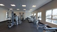 This fitness center offers a well-organized and fully equipped workout environment designed to support a wide range of exercise needs. The space is laid out on a single floor, with a generous open floor plan that provides ample room to move freely between equipment stations.

The cardio area features a row of treadmills and stationary bikes lined along the windowed wall, benefiting from natural light that streams in through large windows. The strength training section is equally well-appointed, with a multi-station cable machine, weight equipment, and additional resistance training apparatus positioned along the opposite wall.

The interior design is clean and functional, with neutral white walls, a dark rubberized flooring ideal for high-traffic athletic use, and a dropped ceiling fitted with recessed fluorescent panel lighting that ensures the space is evenly and brightly illuminated throughout. A flat-screen television is mounted on the wall, adding an entertainment option for users during workouts.

The overall ambiance is modern and professional, reflecting a facility maintained to a high standard. The combination of thoughtful equipment placement, practical finishes, and a bright, airy atmosphere makes this fitness center a well-rounded space suited for both cardio and strength training routines.