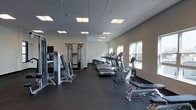 This fitness center offers a well-organized and fully equipped workout environment designed to support a wide range of exercise needs. The space is laid out on a single floor, with a generous open floor plan that provides ample room to move freely between equipment stations.

The cardio area features a row of treadmills and stationary bikes lined along the windowed wall, benefiting from natural light that streams in through large windows. The strength training section is equally well-appointed, with a multi-station cable machine, weight equipment, and additional resistance training apparatus positioned along the opposite wall.

The interior design is clean and functional, with neutral white walls, a dark rubberized flooring ideal for high-traffic athletic use, and a dropped ceiling fitted with recessed fluorescent panel lighting that ensures the space is evenly and brightly illuminated throughout. A flat-screen television is mounted on the wall, adding an entertainment option for users during workouts.

The overall ambiance is modern and professional, reflecting a facility maintained to a high standard. The combination of thoughtful equipment placement, practical finishes, and a bright, airy atmosphere makes this fitness center a well-rounded space suited for both cardio and strength training routines.