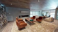 Located at 850 Pacific St in Stamford, Connecticut, this single-floor amenity space combines refined design with functional versatility. The centerpiece of the layout is an expansive lounge area anchored by a sleek, modern fireplace set against a concrete-textured accent wall, above which a framed landscape artwork adds an artistic focal point. A live-edge wood coffee table sits atop a plush, textured area rug, surrounded by cognac leather lounge chairs with polished chrome bases, creating an inviting and upscale atmosphere. The warm, natural wood plank ceiling runs throughout the space, providing a cohesive design element that contrasts beautifully with the cool gray walls. Floor-to-ceiling glass partitions allow abundant natural light to flow through the lounge and offer views into the adjacent fitness area, which is equipped with exercise machines. A dramatic floor-to-ceiling bookshelf installation serves as a striking visual feature along one wall, adding depth and character to the lounge. The space also includes a well-appointed conference and business room, featuring a marble-topped conference table, upholstered chairs, a wall-mounted flat-screen display, built-in shelving, and office equipment — ideal for meetings and productivity. Tile flooring, recessed lighting, and thoughtful spatial flow complete this polished, multi-functional environment.