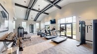 Located at 285 Nance Rd in Huntsville, Alabama, this fitness center is a thoughtfully designed single-floor facility offering a comprehensive workout environment. The space is defined by its dramatic vaulted ceiling adorned with bold, dark cross-beam detailing and modern pendant lighting, creating a striking architectural focal point that enhances the sense of openness and volume throughout.

The cardio zone is well-appointed with multiple treadmills, elliptical trainers, a stationary bike, and a rowing machine, all positioned to take advantage of the abundant natural light pouring in through large floor-to-ceiling windows and glass double doors. These expansive openings provide views of the surrounding outdoor greenery, contributing to an energizing and open atmosphere during workouts.

The strength training area includes a cable machine, a multi-functional weight machine, a full dumbbell rack with a range of weights, and adjustable benches — providing options for both beginners and experienced athletes. Large wall-mounted mirrors in the strength zone enhance spatial awareness and allow users to monitor their form. A wall-mounted flat-screen TV is prominently placed for entertainment during cardio sessions.

The flooring consists of durable, textured carpet tiles that define different workout zones within the space. The neutral color palette of light gray walls paired with dark trim and equipment gives the facility a clean, modern aesthetic suited to a focused and motivating exercise environment.