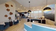 Located at 7660 S Winnipeg St in Aurora, Colorado, this single-floor community clubhouse offers a thoughtfully designed, open-concept space that seamlessly blends functionality with modern style. The layout flows naturally from a welcoming reception area — anchored by a curved, teal-accented granite-topped desk with pendant lighting — into a spacious lounge and communal gathering zone.

The lounge is a standout feature, boasting soaring ceilings, light hardwood-style flooring, and a dramatic large-scale mountain mural that serves as a bold focal point. Comfortable seating arrangements include plush sofas, leather accent chairs, and layered area rugs, all complemented by statement lighting including a candle-style chandelier and contemporary dome pendants. Natural light fills the space through tall windows with slatted blinds.

The fully equipped kitchen area features dark cabinetry contrasted against steel-blue accent walls, stainless steel appliances including a French door refrigerator and range, and an expansive granite island with bar seating — ideal for community events. An adjacent dining area with a wall-mounted TV and map-print artwork adds a social gathering space.

Throughout the clubhouse, the design palette of warm neutrals, teal, and deep blue creates a cohesive, inviting atmosphere suited for both casual use and organized community activities.
