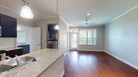 This single-floor residence offers a thoughtfully designed open-concept layout that seamlessly connects the kitchen and living areas. Rich hardwood flooring flows throughout the main living spaces, complemented by crown molding that adds a refined architectural touch. The kitchen is well-equipped with dark espresso cabinetry, granite countertops, stainless steel appliances including a refrigerator, electric range, and built-in microwave, all set against a classic tile backsplash. A peninsula counter extends the workspace and provides a natural transition into the open living area, which is bathed in natural light from a large window and glass door leading to an outdoor balcony. The ceiling fan overhead adds both function and comfort to the living space. The bedroom features plush carpeting, an arched window with views of the surrounding neighborhood, and generous proportions. The bathroom is finished with tile flooring, dark vanity cabinetry, granite countertops, and a large mirror. A spacious walk-in closet with built-in shelving provides ample storage. An additional room with hardwood floors and a built-in desk nook offers versatile functionality. Overall, the space presents a clean, neutral palette throughout.