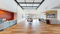 This generously sized single-floor communal space offers a sophisticated, open-concept layout that seamlessly integrates a living room, dining area, and kitchen into one cohesive environment. Wide-plank hardwood-style flooring flows throughout, anchoring the clean, modern aesthetic defined by crisp white walls and bold architectural accents.

The main gathering area is a standout, featuring a striking glass-panel ceiling structure framed in black steel that floods the space with natural light. A substantial live-edge wood dining table anchors the center of the room beneath a cluster of statement pendant lights. Large black-framed windows line the perimeter, offering views of a resort-style pool surrounded by palm trees, reinforcing the bright and airy atmosphere.

The kitchen area features light sage cabinetry paired with a bold red subway tile accent wall, stainless steel appliances, and a beverage station, providing both functionality and a vibrant pop of color. A bar-height counter with seating offers an additional casual dining option.

The living area transitions into a dedicated game room, complete with a pool table, a large multi-panel display screen, and textured copper-toned wall panels that lend an industrial warmth. Slatted wood paneling and carefully curated artwork are woven throughout, adding layers of texture and visual interest. The overall ambiance is modern, energetic, and thoughtfully designed for social engagement.