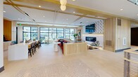Located at 2501 NJ-37 E in Manchester, New Jersey, this contemporary two-story clubhouse is a thoughtfully designed community amenity space that blends sophisticated aesthetics with practical functionality. The upper level commands attention with soaring vaulted ceilings, dramatic exposed steel beams, and large black-framed windows that flood the space with natural light and offer views of the surrounding neighborhood. This expansive floor features an open-concept lounge and social area with a full kitchen outfitted with light wood cabinetry, a white quartz island with bar seating, and direct outdoor access. A dedicated game room on this level showcases a striking sculptural ceiling with layered brass and globe fixtures, complemented by a poker table, shuffleboard, and glass-partitioned walls. Four private offices provide quiet, professional workspaces, while cozy lounge nooks and a formal conference room with woven accent chairs serve a variety of gathering needs. The lower level serves as the welcoming entry point, featuring an elevator, staircase access, and a refined reception area adorned with large-scale abstract artwork, gold-accented beam details, and curated furnishings. Throughout both floors, a cohesive design palette of warm wood tones, neutral stone-look tile flooring, and modern globe lighting creates an inviting and polished ambiance.