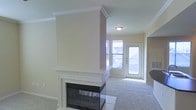 This single-floor space is centered around a striking dual-sided fireplace, which serves as a architectural focal point dividing the living area. The fireplace features a classic white mantel with crown molding detail, a black firebox surround, and a tile hearth, adding both functionality and visual interest to the space.

The living area is finished with wall-to-wall carpeting in a neutral tone, complementing the warm beige walls throughout. Crown molding runs along the ceiling, lending a refined touch to the interior. A flush-mount ceiling light provides overhead illumination, while natural light filters in generously through a large window and a glass-paneled entry door near the front of the space.

The entryway is bright and welcoming, benefiting from dual windows that allow daylight to flood the area. A glimpse of an adjacent room — likely a kitchen or dining area — is visible, suggesting an open and connected layout. The overall color palette of creams and neutrals creates a clean, neutral canvas throughout the home, making it feel bright, open, and move-in ready.