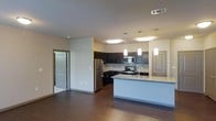 This single-floor space showcases a thoughtfully designed open-concept layout that seamlessly connects the kitchen and living areas. The kitchen is a standout feature, outfitted with dark espresso cabinetry that extends to the upper and lower walls, providing ample storage. Stainless steel appliances — including a refrigerator, range, and built-in microwave — complement the sleek aesthetic. A large kitchen island with light granite-look countertops offers generous prep space and serves as a natural gathering point. Three pendant lights hang above the island, adding focused illumination and a contemporary design touch, while recessed flush-mount ceiling lights brighten the rest of the space. A tile backsplash adds texture behind the range area. The dark hardwood-style flooring runs throughout, creating a cohesive and grounded feel. Neutral wall tones keep the space bright and versatile. Doorways visible from the main area suggest additional rooms beyond, and a paneled interior door hints at a well-appointed layout. Overall, the space conveys a clean, modern aesthetic with functional design throughout.