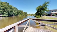Located in Charleston, South Carolina, Castlewood Townhomes offers a peaceful community environment highlighted by a standout natural amenity: a wooden dock extending over a serene, expansive pond. The dock is constructed with sturdy wooden planking and cross-braced railings, providing a safe and inviting space to take in the surrounding scenery. The pond itself is a central feature of the community, its calm waters reflecting the brilliant blue skies and the dense canopy of mature trees that frame the shoreline. The lush, verdant tree line encircling the pond creates a sense of seclusion and natural beauty, offering residents a tranquil escape from the everyday. Views from the dock extend across the water to the opposite bank, where additional greenery and glimpses of neighboring structures can be seen, reinforcing the cohesive, nature-integrated character of the community. The single-level layout of the community ensures easy accessibility throughout. Whether used for quiet reflection, casual recreation, or simply enjoying the outdoors, this waterfront dock and pond area serves as a defining feature of the Castlewood Townhomes community in West Ashley, Charleston.