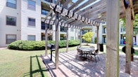 This community amenity space offers a thoughtfully designed outdoor environment ideal for gathering and leisure. At its centerpiece is a charming courtyard featuring a large wooden pergola structure that provides partial shade over a brick-paved patio. The pergola area is equipped with metal table-and-chair seating and a charcoal grill, making it well-suited for outdoor dining and casual cookouts. The surrounding grounds are lushly landscaped with manicured shrubs, ornamental trees, and open green lawn areas, all framed by the multi-story residential buildings that enclose the courtyard.

The space also includes a generously sized covered lounge pavilion that opens to the outdoors through wide arched and open-frame openings, allowing for abundant natural light and refreshing breezes. This covered area is furnished with bold red-cushioned armchairs and a dark wood coffee table, complemented by a wall-mounted flat-screen television and a vending machine for added convenience. An additional covered porch area features wicker seating with vibrant floral cushions and decorative wall art, lending a warm and welcoming atmosphere. Together, these spaces create a versatile outdoor community hub designed for comfort, convenience, and connection.
