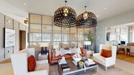 Located at 500 Dalton Way in Little River, South Carolina, this thoughtfully designed single-story space offers a cohesive coastal-contemporary aesthetic across its well-appointed rooms. The open-plan living, dining, and kitchen area serves as the heart of the space, where each zone flows seamlessly into the next. The living area makes a striking impression with oversized woven dome pendant lights suspended above a rich cognac leather sofa, complemented by neutral armchairs and a curated coffee table arrangement. A floor-to-ceiling grid-paned window wall bathes the seating area in warm, diffused light while visually expanding the space. Large framed coastal artwork and paneled gray accent walls add architectural interest and a refined sense of place. The kitchen features generous countertop space with sleek dark stone surfaces, white cabinetry with gold hardware, a full-size stainless steel refrigerator, and a substantial wood-topped island. Woven pendant lights echo the living area's coastal motif. A bright office or workspace area includes large windows overlooking the surrounding greenery, providing an airy, productive environment with warm hardwood flooring throughout. The hallway connects the areas gracefully, lined with curated artwork and sconce lighting, reinforcing the space's polished, detail-oriented character.