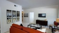 This single-floor space centers around a well-appointed living area that combines comfort with a clean, contemporary aesthetic. The room is finished with light neutral walls and pale carpeting, creating a bright, airy atmosphere that feels open and inviting. Crown molding adds a refined architectural detail, lending a polished character to the space.

The living area is anchored by a bold rust-orange sofa paired with a sleek glass-top coffee table, offering both style and practicality. A wall-mounted flat-screen television sits above a dark wood media console, providing a natural focal point for the room. Decorative accents, including a pair of large birch tree canvas paintings, contribute a nature-inspired artistic touch to the otherwise minimalist décor.

A white six-panel door leads to an adjacent room, suggesting a thoughtfully connected floor plan. A glimpse into the hallway reveals additional living areas, hinting at further functional spaces within the home. The overall ambiance is calm and well-organized, with tasteful staging that highlights the versatility and livability of this single-story residence.