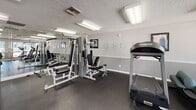 This fitness center is a compact yet well-appointed exercise facility designed to support a range of workout routines. The space is finished with durable dark rubber flooring throughout, providing a practical and slip-resistant surface suited for active use. Textured ceilings with flush fluorescent lighting ensure consistent, bright illumination across the room.

The cardio area is equipped with multiple Landice treadmills, elliptical trainers, and a recumbent stationary bike, offering a variety of low-impact and high-intensity options. A Hoist multi-station cable and weight machine anchors the strength training section, complemented by a large wall-mounted mirror that visually expands the space and aids in form monitoring during exercise.

Large windows along one wall allow natural light to filter in while providing views of a landscaped outdoor pool area, creating an open and energizing atmosphere. A built-in reception or check-in counter is positioned near the entrance, adding a structured, organized feel to the space. Posted signage and framed artwork are visible along the walls, contributing to a clean, professional environment. The facility is accessed through a dedicated doorway, maintaining separation from adjacent areas.
