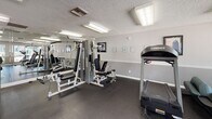 This fitness center is a compact yet well-appointed exercise facility designed to support a range of workout routines. The space is finished with durable dark rubber flooring throughout, providing a practical and slip-resistant surface suited for active use. Textured ceilings with flush fluorescent lighting ensure consistent, bright illumination across the room.

The cardio area is equipped with multiple Landice treadmills, elliptical trainers, and a recumbent stationary bike, offering a variety of low-impact and high-intensity options. A Hoist multi-station cable and weight machine anchors the strength training section, complemented by a large wall-mounted mirror that visually expands the space and aids in form monitoring during exercise.

Large windows along one wall allow natural light to filter in while providing views of a landscaped outdoor pool area, creating an open and energizing atmosphere. A built-in reception or check-in counter is positioned near the entrance, adding a structured, organized feel to the space. Posted signage and framed artwork are visible along the walls, contributing to a clean, professional environment. The facility is accessed through a dedicated doorway, maintaining separation from adjacent areas.